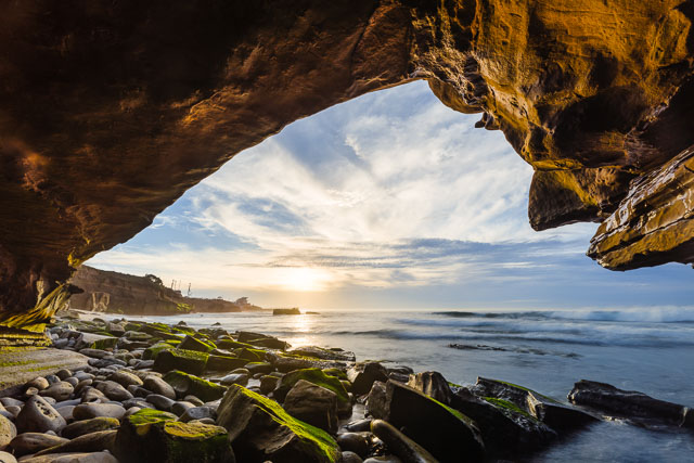 A seascape seen through the opening of a cave on the La Jolla coast. The sky is a pastel blue with white clouds. The shoreline is rocky with some covered in green algae. The tide is coming in and waves can be seen in the ocean.