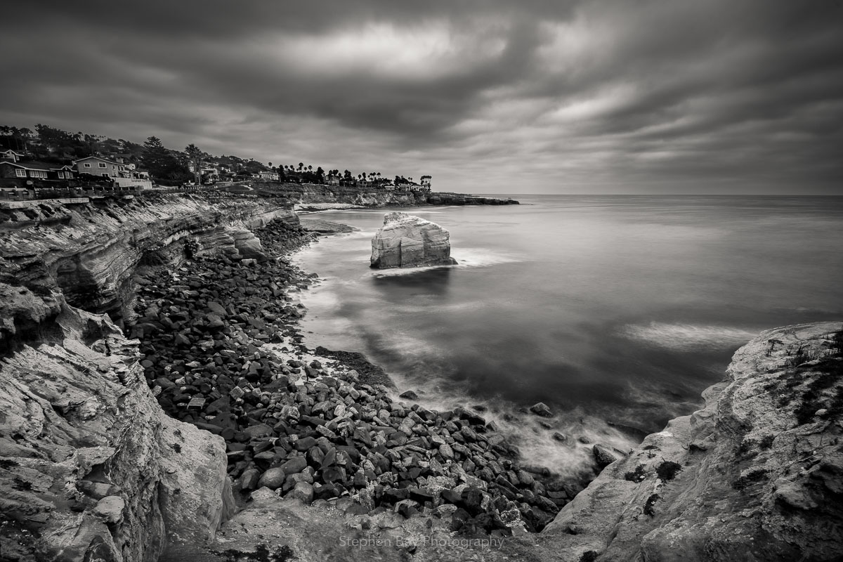 A black and white photo of the coastline at sunset cliffs. The coastline starts at the viewers feet and then curves around into the distance. In the mid-ground is Ross Rock.