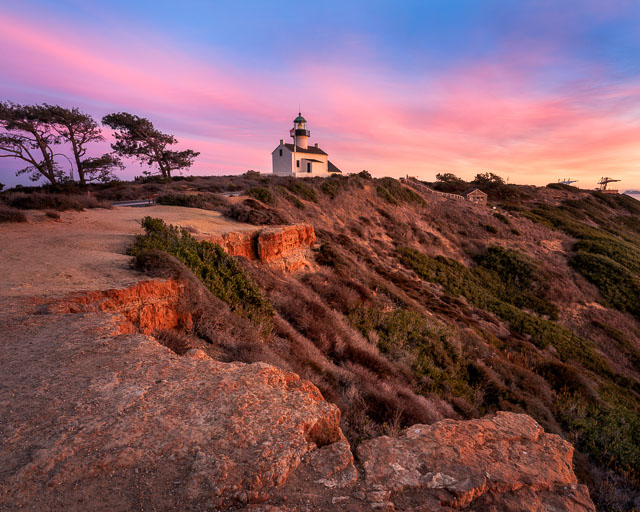 The old Point Loma lighthouse at sunset set against a rugged cliff edge.