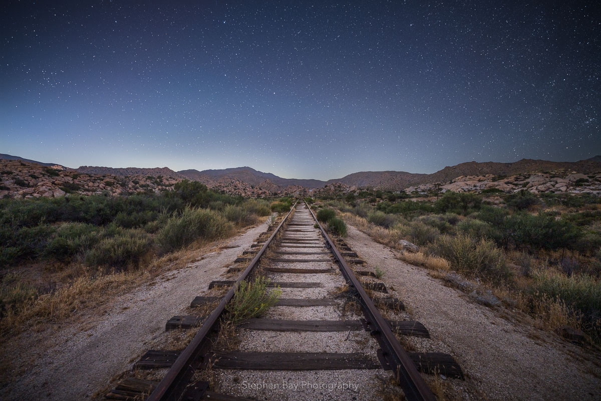 Train tracks in the center of the frame lead off into the desert and distant mountains. The night sky is full of stars.