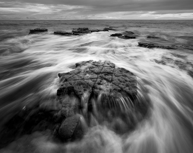 Water rushing over the rocks on a beach at Sunset Cliffs. The photo is in black and white.