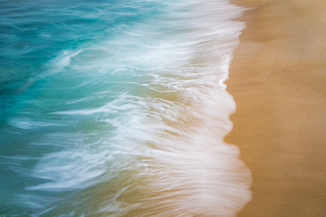 A close-up of ocean waves gently flowing up onto a sandy beach. The water is blue-green or turquoise/aqua in color with white highlights.