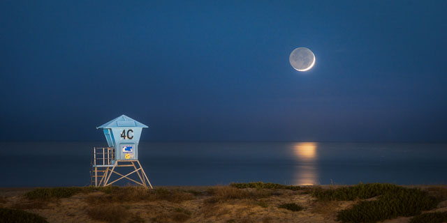 A waxing crescent moon sets over a lifeguard tower and beach. The scene is at night with dark blue skies and the moon is reflecting into the water. The unlit portion of the moon can be seen. This is called earthshine. The image is panorama format