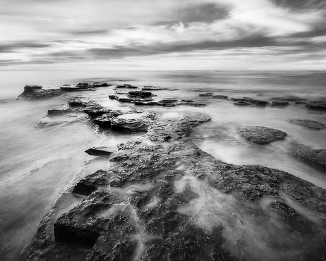A black & white photo of a rock formation at Sunset Cliffs, San Diego. The rock formation has sharp jagged edges and ocean water is flowing over the top