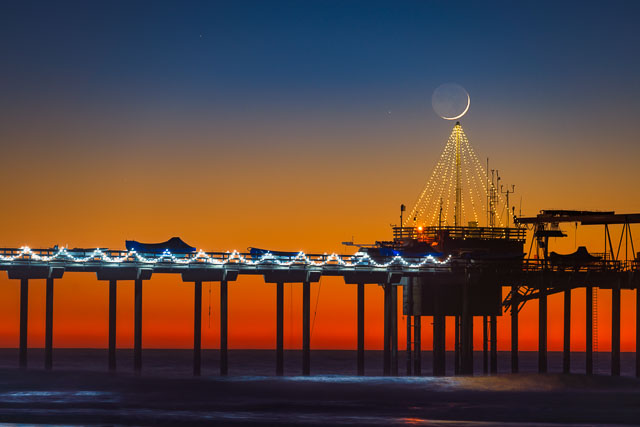 A crescent moon setting over the Christmas Tree at Scripps Pier. The moon is just above the top of the tree.