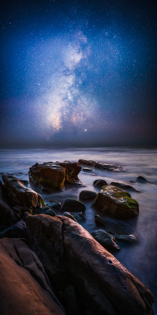 A vertical panorama of the coastline at Windansea Beach in La Jolla at night. The foreground has a number of sandstone rocks with the ocean water swirling around. The sky shows the stars of the Milky Way galaxy and the central core.