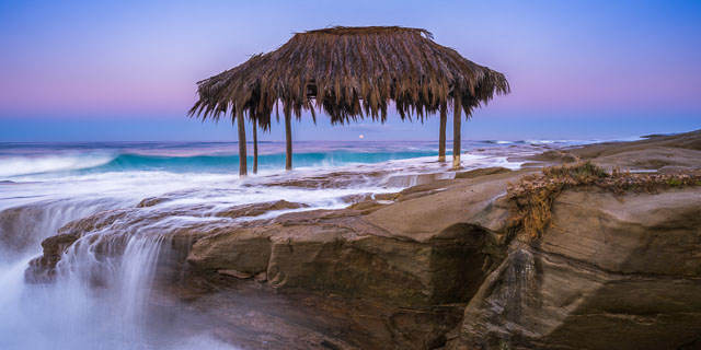Panorama of the Windansea Beach hut in La Jolla just before sunrise. The sky is a  rich blue/magenta and the full moon is setting in the distance. The water from the ocean waves has come up onto the rocks.
