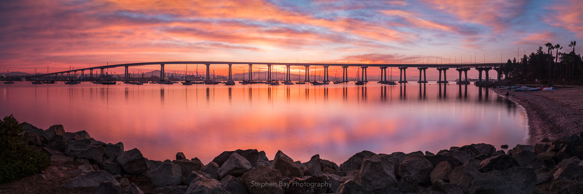 Coronado Sunrise is a panoramic photo of the Coronado Bridge at sunrise by San Diego photographer Stephen Bay. The artwork shows the entire span of the bridge set against a dramatic orange and pink sky which is reflected into the water.
