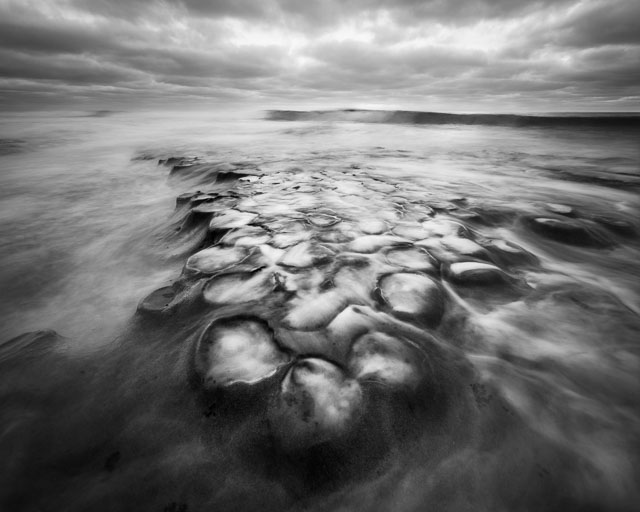 A black and white photo of the potholes at Hospitals reef in La Jolla. The water is swirling in the little craters and appears as if the water is boiling.