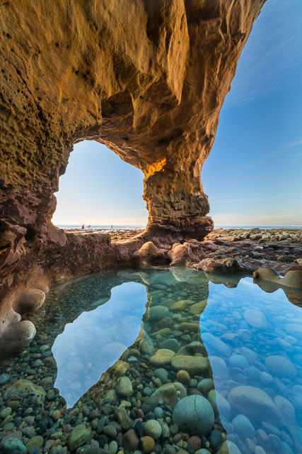 In the foreground is a tide pool with turquoise or aqua water. There is a red sandstone cliff with an opening or window formed by erosion. The blue sky reflects into the tide pool.