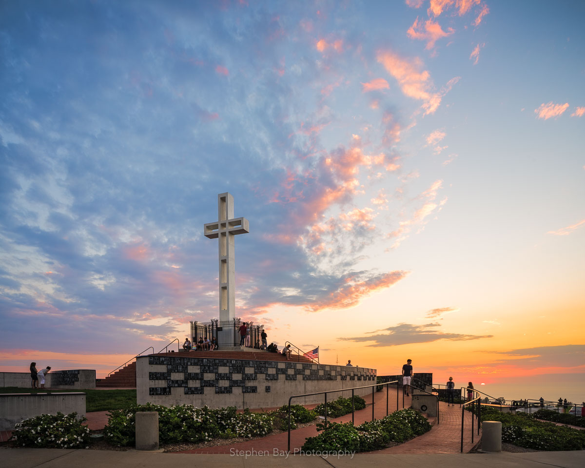 The Mt Soledad Veteran's Memorial in La Jolla. The cross is set against a light blue sky just before sunset. The sun is about to disappear from the horizon and is just barely able to light up the edge of the clouds in the sky.