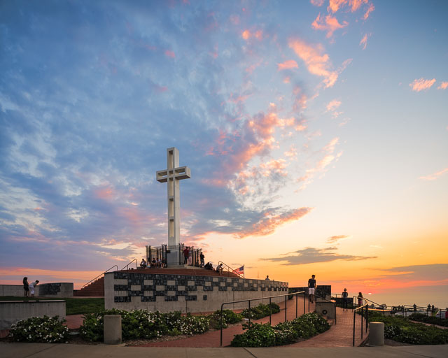 The Mt Soledad Veteran's Memorial in La Jolla. The cross is set against a light blue sky just before sunset. The sun is about to disappear from the horizon and is just barely able to light up the edge of the clouds in the sky.