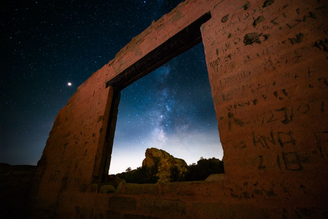 The remains of an abandoned house with only wall left standing with an opening for a window. In the center of the window the core of the Milky Way can be seen.