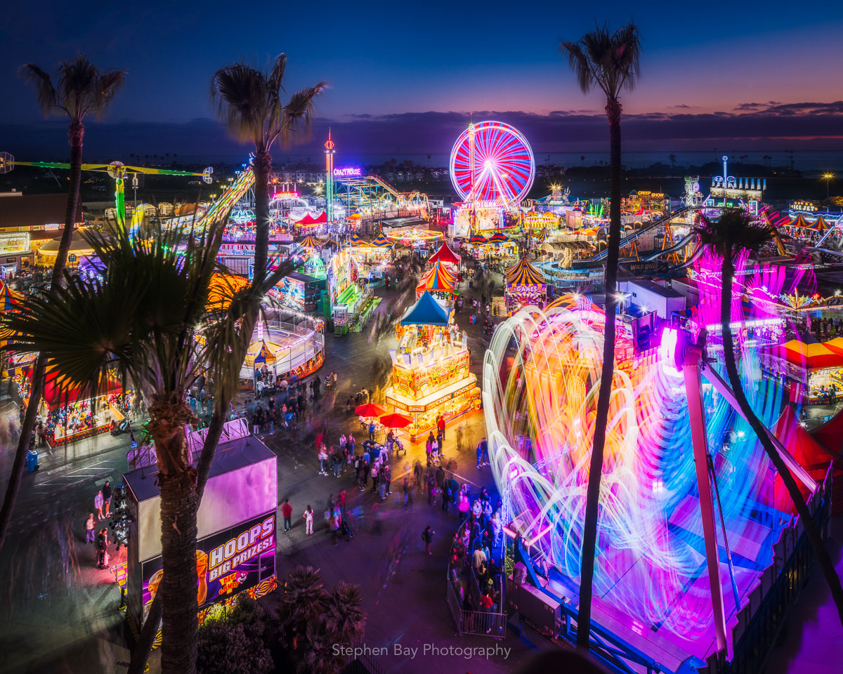 Looking down on the fun zone of the san diego fair where there are multiple amusement park rides. The rides are in operation and leave bright colorful light trails as they move during a long exposure. Crowds of people can be seen moving about and waiting in line