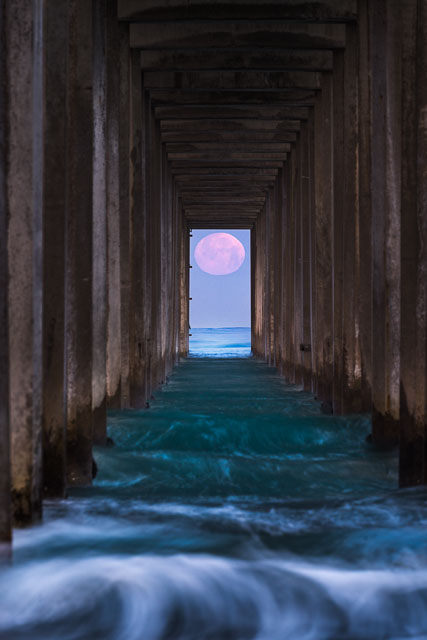 The full moon is centered in the opening between the support pylons at scripps pier.