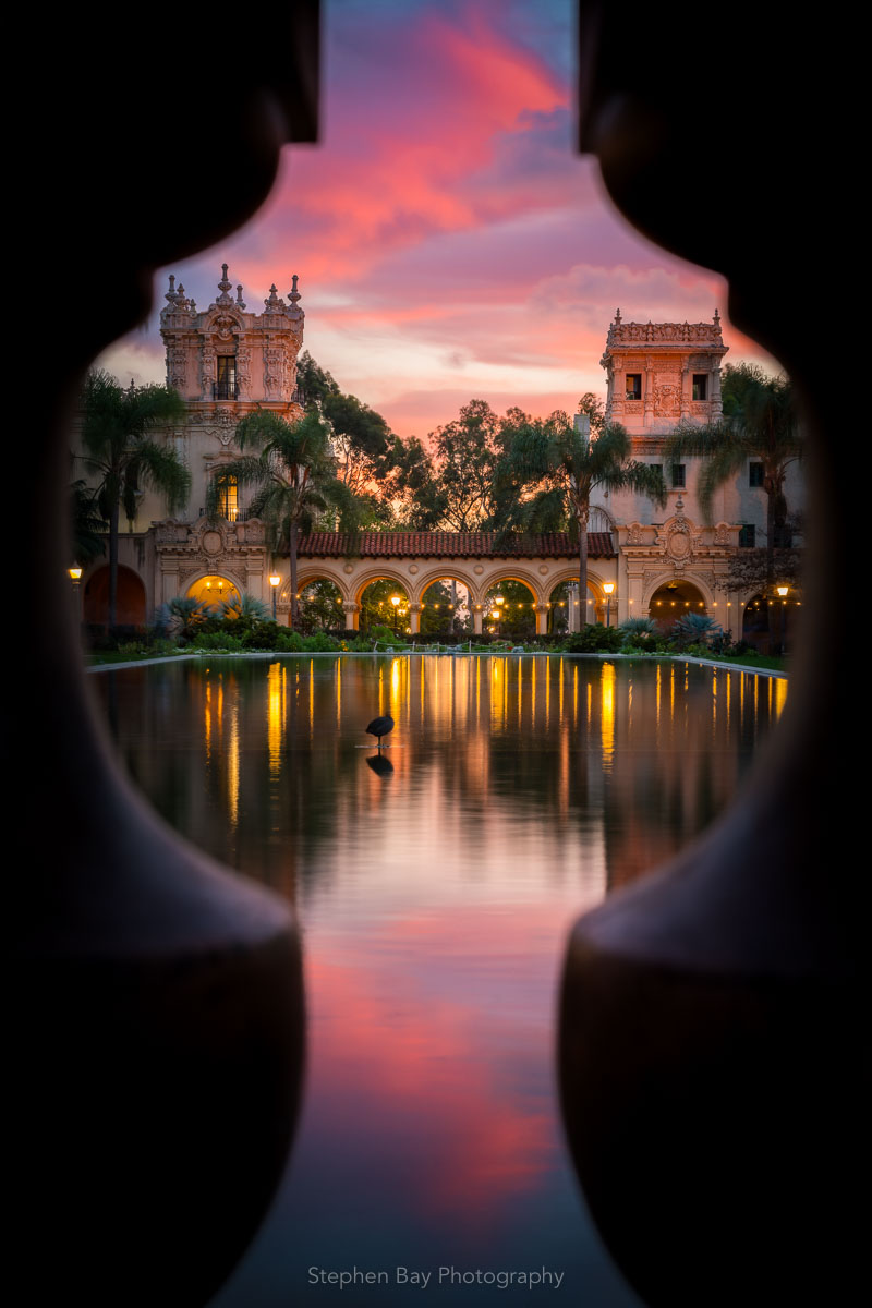 Enchanted is a vertical photo of the Lily pond in Balboa Park by San Diego photographer Stephen Bay. The artwork shows the pond through the balusters on a small bridge creating a keyhole view. The sun has just set and fills the sky with pink and magenta tones which reflects into the pond. Casa de Balboa and the House of Hospitality can be seen at the far end of the pond.