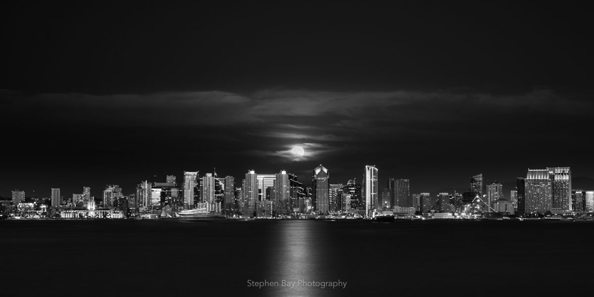 The full moon rising over the San Diego skyline from across the bay. The moon is just above the tops of the buildings with a dark black sky above that. 