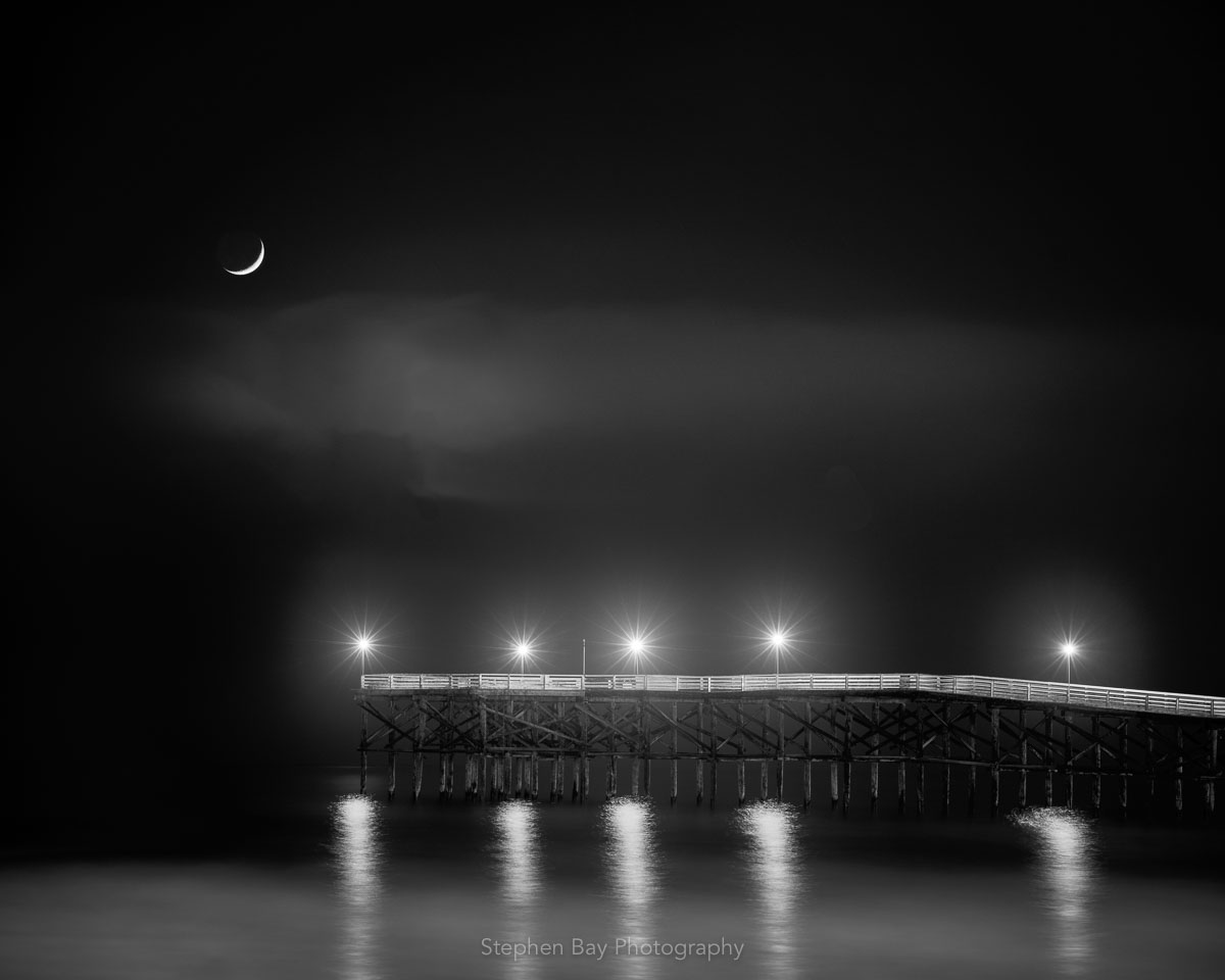 A photo of the crescent moon setting on the Crystal Pier at Pacific Beach, San Diego. The photo is black and white and you can see the reflections of the pier lights in the ocean water.