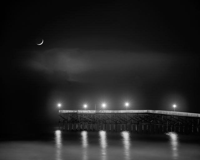 The crescent moon setting on the Crystal Pier at Pacific Beach, San Diego. The photo is black and white and you can see the reflections of the pier lights in the ocean water.