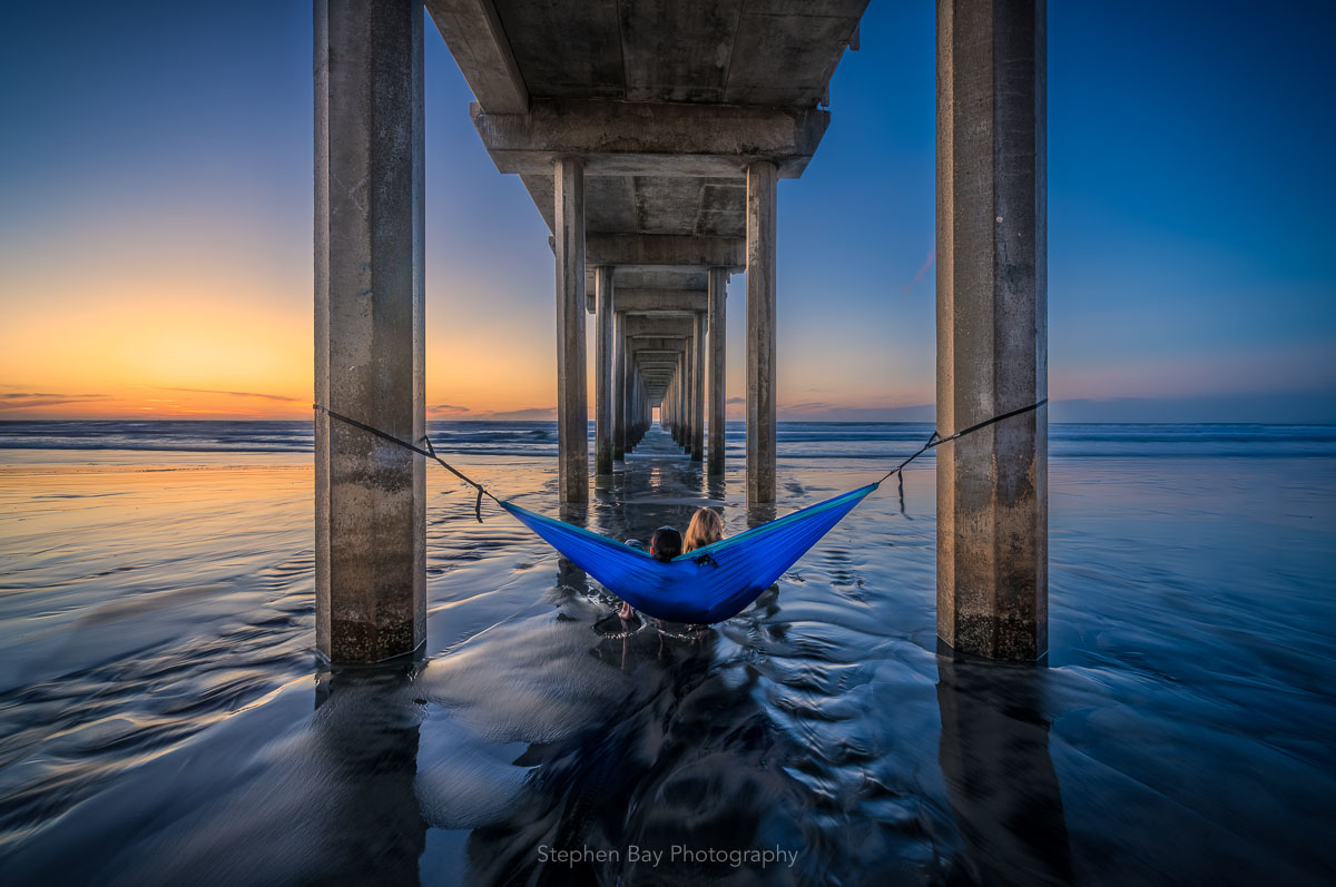 My Happy Place is a photo of a couple in a hammock at Scripps Pier in La Jolla. The artwork represents peace and relaxation, and has predominantly blue tones with an orange glow on the horizon as the sun sets. This photo is by Stephen Bay, a San Diego photographer.