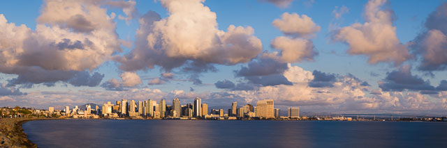 A panorama of the San Diego skyline with many cumulus clouds floating in over the bay. The photo was taken from Harbor Island in the late afternoon. On the right you can see the Coronado Bridge.