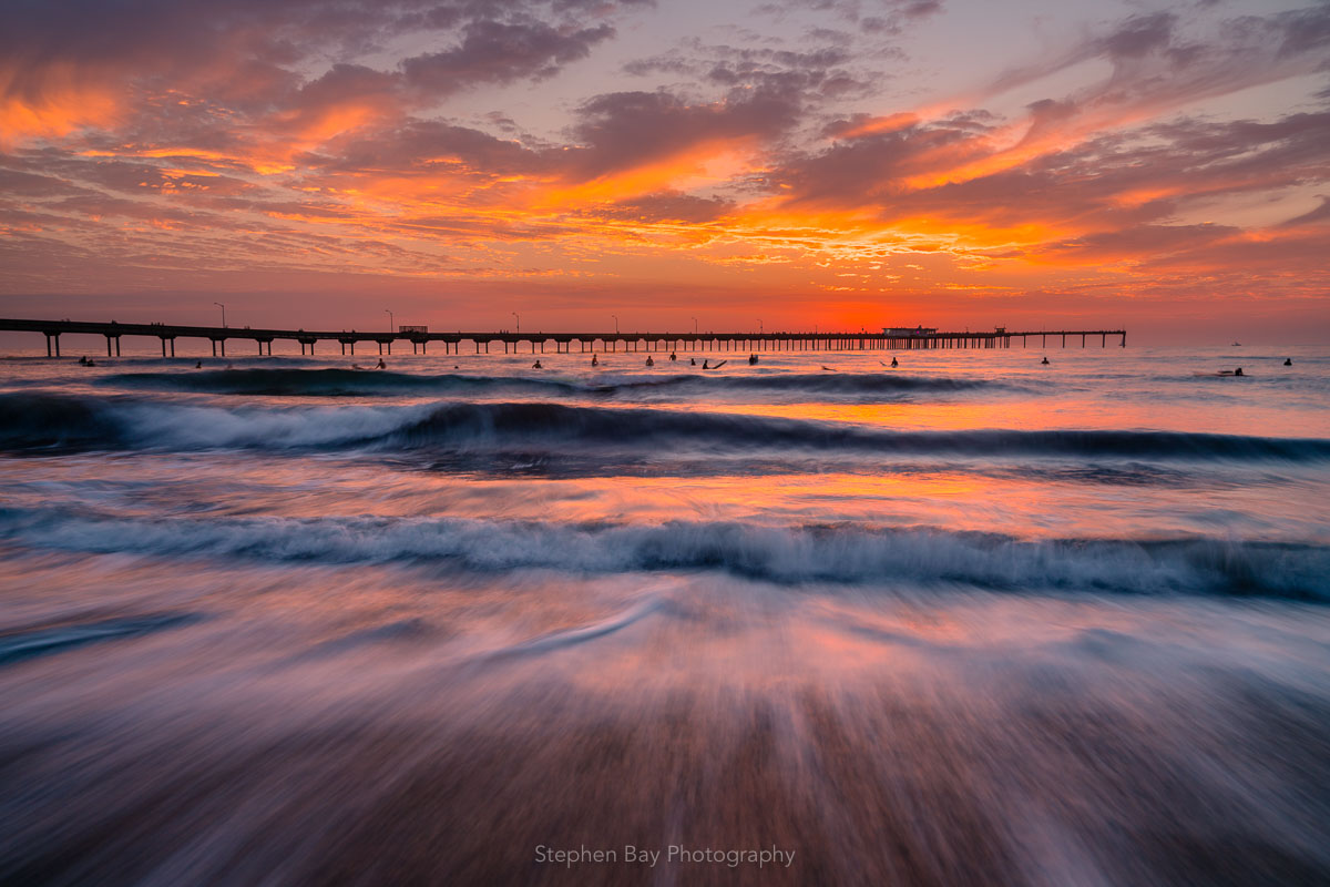 Endless Waves is a landscape photograph by Stephen Bay. It depicts a multitude of waves coming ashore with surfers and the Ocean Beach Pier in the background. The sky is predominately orange from a dramatic sunset and this light is reflected in the water. The viewpoint of the artwork is from a person standing within the water as waves come up the beach.