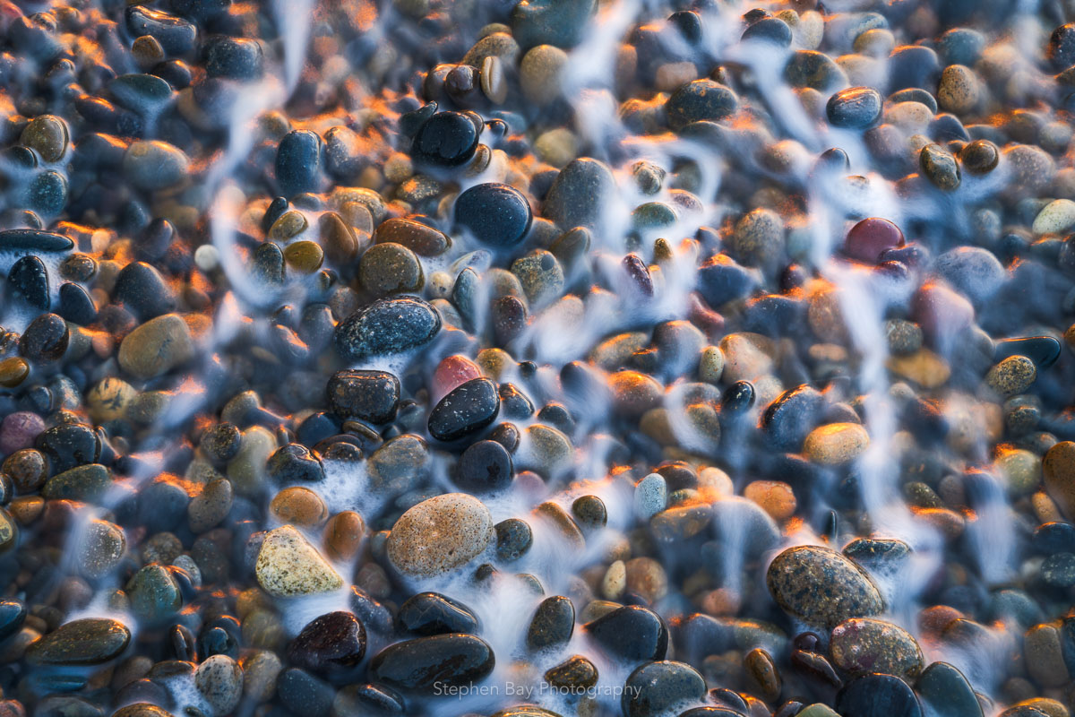 A close up of small round pebbles on the beach. This is an overhead view and water is running back into the ocean leaving a misty trail.