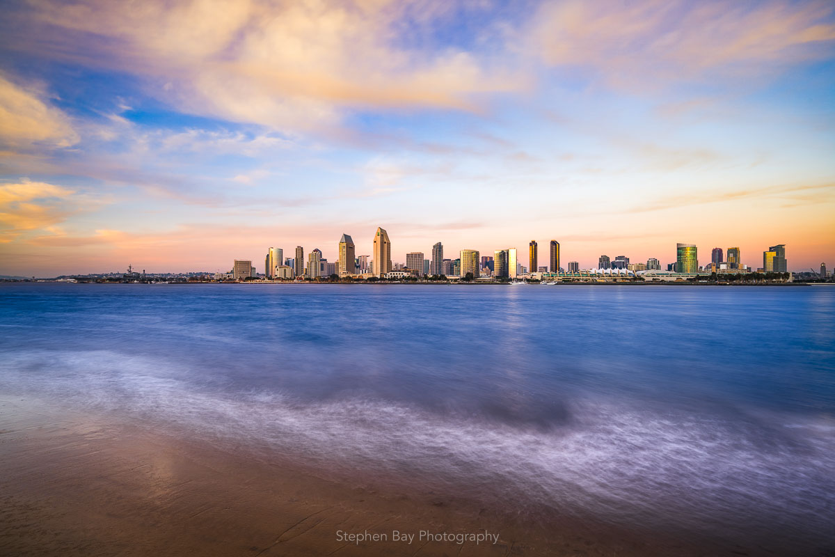 A photo of the San Diego skyline. There are clouds blowing in from offshore that give the image a dream feel. The waves are washing up against the sandy shoreline.
