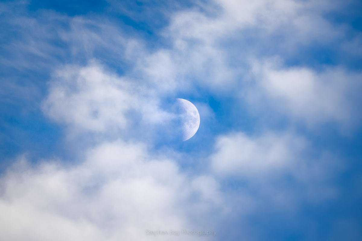 The first quarter moon amongst the clouds in a blue sky. The moon is a half circle and surrounded by white clouds. No land is visible.