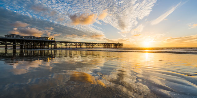 Panorama of the crystal pier at Pacific Beach, San Diego just before sunset. The sun is low on the horizon and casting warm rays of light on the clouds which is reflected onto the wet sand.