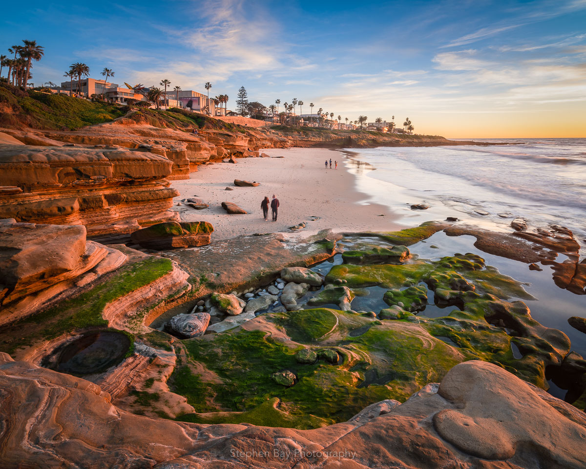 The La Jolla coastline with exposed rocks covered in green algae. There is warm light from the setting sun. In the distance, there is a couple walking on the beach enjoying the moment.