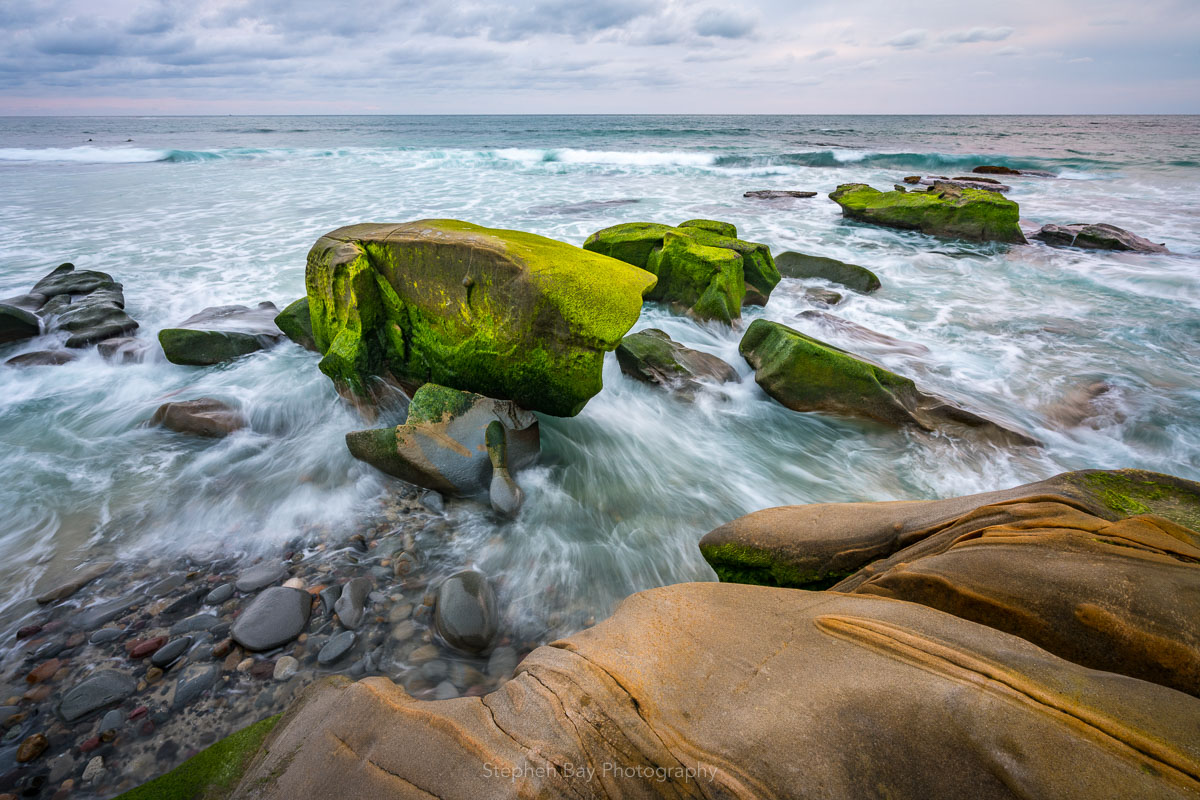 Green algae covered rocks at Windansea beach. The sand has been washed away by winter storms and the waves and water are rushing in at the base of the rocks. The sky is overcast.