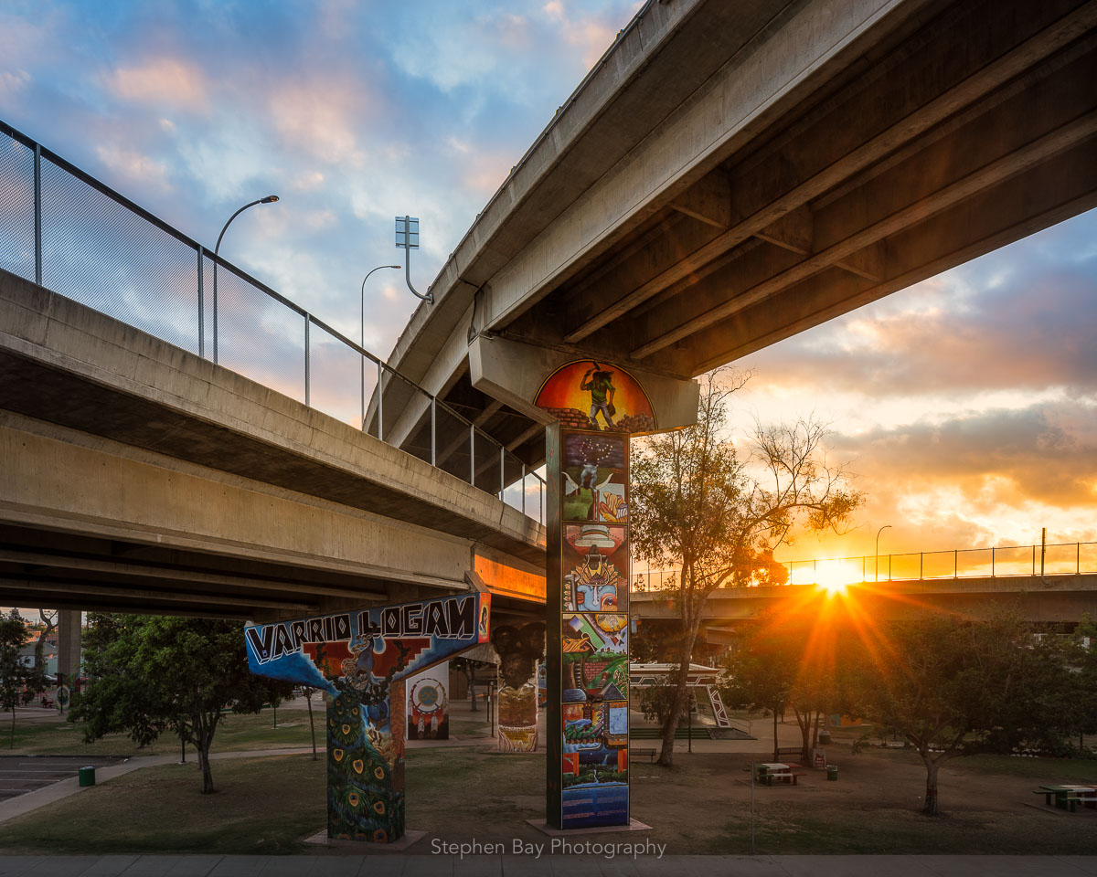 A photo of Chicano Park which is in Barrio Logan. The park is situated underneath the ramps to the Coronado Bridge and you can see concrete pillars and the on and off ramps. The pillars are adorned by murals and there is a stage in the middle of the park. The sun is setting and casts a warm glow on everything.